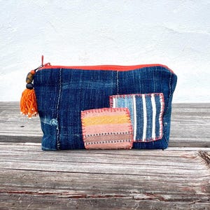 Puede incluir: Una bolsa de mezclilla azul con cremallera y borla naranja. La bolsa presenta tres parches de tela coloridos con bordes cosidos. Los parches incluyen rayas y patrones geométricos. La bolsa está sobre una superficie de madera.