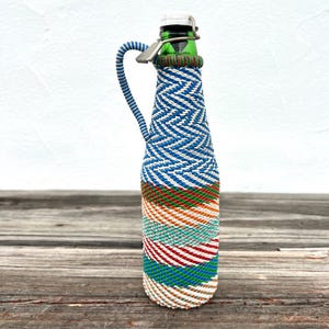 Könnte beinhalten: Eine dekorative Glasflasche mit gewebter Außenseite. Die Flasche ist mit einem blau-weißen Zickzackmuster umwickelt, mit roten, orangefarbenen, grünen und weißen Bändern. Sie hat einen Metallverschluss und einen blauen Griff. Die Flasche ist etwa 25 cm hoch.