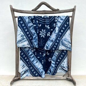 May include: A rectangular textile featuring a deep indigo blue and white tie-dye pattern with floral motifs and geometric bands. The fabric is draped over a rustic, weathered wooden frame with a curved top bar and four vertical support legs.