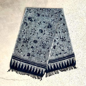 Könnte beinhalten: Ein blau-weiß gemusterter Schal mit floralem Design. Der Schal hat einen dunkelblauen Rand mit Fransenbesatz. Der Schal ist zur Hälfte gefaltet und liegt auf einer hellen Oberfläche.