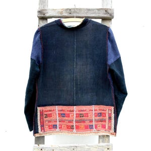 Peut inclure: Veste bleu foncé avec des manches en patchwork bleu et vert. L'ourlet inférieur présente un panneau décoratif brodé rouge et blanc. La veste est suspendue à une échelle en bois sur fond blanc. La veste a un col rond.