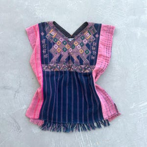 Op de afbeelding: Een mouwloze tuniek met een donkerblauw middenpaneel met verticale strepen en een V-hals. Het bovenste gedeelte toont ingewikkeld geometrisch borduurwerk in roze, groen en geel. De zijkanten en de zoom zijn roze met franjes als afwerking.