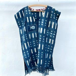 Op de afbeelding: Een mouwloze, indigoblauwe tuniek met een V-hals en een wit verticaal streepjespatroon. Een kleine vierkante patch met een bladerontwerp is op de voorkant genaaid. De onderkant heeft een franje detail. De tuniek hangt aan een houten hanger.