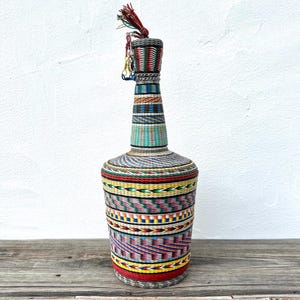 May include: A tall, decorative bottle wrapped in woven, colorful fibers. The bottle features a cylindrical base and a long, slender neck, adorned with bands of red, yellow, blue, and green. A tassel hangs from the top.