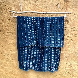 Könnte beinhalten: Indigoblaues Textil mit weißem Punktmuster, aufgehängt an einer natürlichen Holzstange. Der Stoff ist gefaltet, wodurch die vertikalen Linien des Musters und die Textur des Materials sichtbar werden. Hintergrund: eine strukturierte, hellbraune Wand.