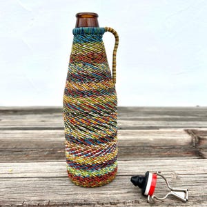 May include: A decorative bottle with a brown neck and a colorful woven exterior. The bottle has a handle and is next to a bottle stopper with a black top, red and white accents, and a silver metal opener.