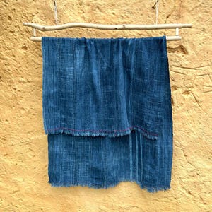 Peut inclure: Un textile bleu indigo foncé suspendu à une tringle en bois naturel. Le tissu a un aspect texturé avec des lignes verticales et un bord effiloché avec un fin accent rouge. Le textile est présenté contre un mur texturé de couleur jaune.