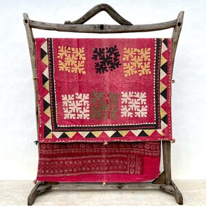 May include: A vibrant red textile with geometric patterns, displayed on a rustic wooden frame. The textile features embroidered squares in gold, black, and white, with a detailed border of triangles. The lower section has a repeating circular design.