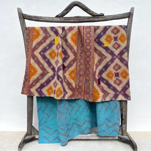 Op de afbeelding: Een gelaagde textielpresentatie met een bovenste quilt met een paars en oranje ruitpatroon en een onderste quilt met een turquoise zigzagontwerp. Beide stukken hangen over een rustiek, verweerd houten frame met een gebogen bovenbalk.