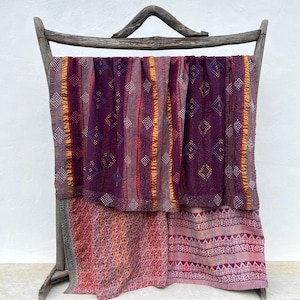 Könnte beinhalten: Ein rechteckiges Textil mit einem tiefvioletten oberen Bereich mit vertikalen Streifen und Rautenmustern sowie einem unteren Bereich mit rosa, orangefarbenen und violetten geometrischen und floralen Motiven, drapiert über einem rustikalen Holzrahmen.