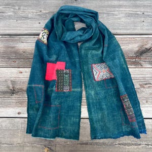 Puede incluir: Bufanda de color turquesa con parches decorativos. La bufanda presenta parches rojos y bordados con varios patrones geométricos. Los parches están cosidos a la bufanda con hilo rojo. La bufanda está hecha de una tela suave y fluida.