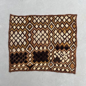 Könnte beinhalten: Rechteckiger Textil mit einem geometrischen Rautenmuster in Braun-, Beige- und Schwarztönen. Die Kanten sind mit einem dunkleren braunen Rand versehen. Das Design zeigt sich wiederholende Rautenformen.
