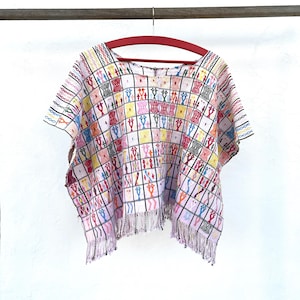 Peut inclure: Un haut blanc à manches courtes présentant un motif à carreaux avec des formes géométriques brodées multicolores en rose, jaune, bleu et orange. Le vêtement possède une encolure ronde et un ourlet fini par de longues franges blanches.