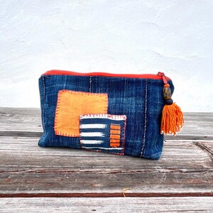 Könnte beinhalten: Eine blaue Jeans-Tasche mit orangefarbenen Aufnähern und einem roten Reißverschluss. Die Tasche hat eine dekorative orangefarbene Quaste mit einer Holzperle. Die Tasche liegt auf einer Holzoberfläche.