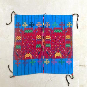 Pode incluir: Um têxtil vibrante com um fundo azul brilhante e bordados vermelhos e multicoloridos intrincados. O design apresenta padrões geométricos e elementos decorativos, com cordões pretos presos nos cantos.