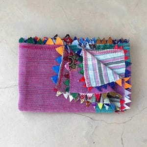Op de afbeelding: Een opgevouwen, magenta gekleurde deken met een patchwork ontwerp. De deken is afgewerkt met kleurrijke driehoekige vlaggen. De binnenkant heeft een gestreept patroon in roze, blauwe en witte tinten. De deken is gemaakt van een textuurstof.