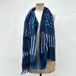 Peut inclure: Écharpe bleu indigo drapée, ornée de motifs géométriques blancs et de franges. Le tissu présente des coutures et des patchs visibles, suggérant une fabrication artisanale. Le design comprend des lignes blanches verticales et pointillées.
