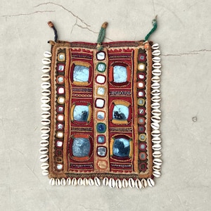 Puede incluir: Textil rectangular hecho a mano con fondo rojo y marrón, con espejos cuadrados y costuras decorativas. Los bordes están forrados con pequeñas conchas blancas. El textil tiene dos lazos en la parte superior para colgar.