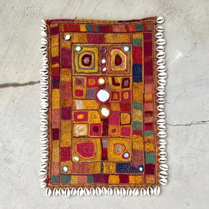 Op de afbeelding: Rechthoekig textiel met een patchwork-ontwerp in tinten rood, geel en oranje. Het centrale patroon heeft vierkanten en rechthoeken met spiegelaccenten. De rand is versierd met witte schelpen.