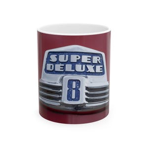 Könnte beinhalten: Weißer Keramikbecher mit roter Außenseite, der ein Vintage-Auto-Emblem-Design aufweist. Das Emblem lautet "SUPER DELUXE 8" in einem Silber- und Blau-Farbschema, das eine klassische Autoästhetik hervorruft.