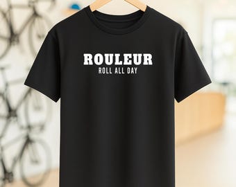 May include: Black crew neck t-shirt with the white text "ROULEUR ROLL ALL DAY". The shirt is hanging on a wooden hanger. The background is blurred, with bicycles visible.