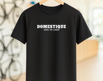May include: Black crew neck t-shirt with the white text "DOMESTIQUE" above "SERVE THE LEADER". The shirt is hanging on a wooden hanger. The background is blurred, with bicycles visible.