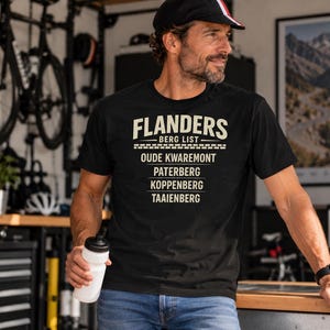 Pode incluir: Uma t-shirt preta de manga curta com texto na cor creme que diz FLANDERS BERG LIST seguido de OUDE KWAREMONT, PATERBERG, KOPPENBERG e TAAIENBERG. O design inclui linhas horizontais e um gráfico com padrão de tijolos.