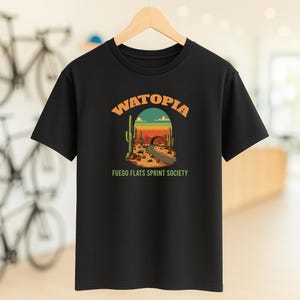 Op de afbeelding: Zwart t-shirt met een woestijnlandschap-afbeelding. Het ontwerp toont een weg, cactussen en de tekst "WATOPÍA" boven "FUEGO FLATS SPRINT SOCIETY" in een boog. Het t-shirt hangt aan een houten hanger.