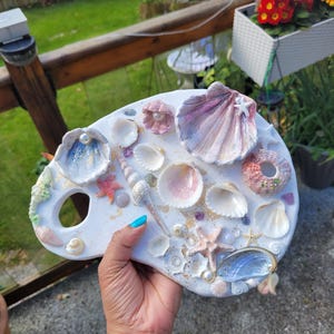 May include: A white palette decorated with seashells, starfish, and pearls. The shells are painted in pink, blue, and white, with gold detailing. The palette is held in a hand, showcasing the seaside theme.