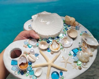 May include: A white, artist's palette-shaped decorative piece adorned with seashells, starfish, and glass beads. The palette features a large scallop shell, various smaller shells, and a starfish. Blue, green, and clear glass beads and pearls add color and texture.