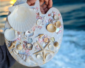 May include: A decorative artist's palette, adorned with various seashells, a starfish, and iridescent gems. The palette is held against a backdrop of a beach at sunset, with the ocean waves in the background. The palette is painted in light blue and gold.