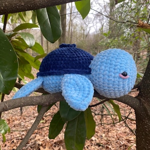 May include: A blue and navy crocheted turtle plush toy sitting on a tree branch. The turtle has a blue shell and a navy blue head and feet.
