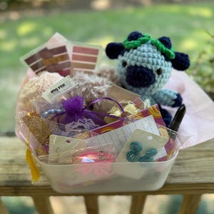 May include: A white rectangular basket filled with various items, including a blue crochet cow, a pen with the text "HUG PEN", and several small wrapped gifts. The basket is lined with pink tissue paper and sits on a wooden deck.