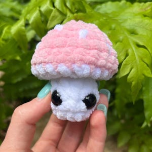 May include: A handmade crochet mushroom toy with a pink cap dotted with white spots and a white stem. The mushroom has large black eyes, giving it a whimsical appearance. The toy is held against a backdrop of green foliage.