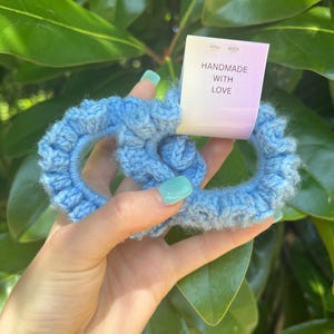 May include: A light blue crocheted hair scrunchie in a pretzel shape. The scrunchie is handmade and has a tag that reads "HANDMADE WITH LOVE". The background features green leaves.