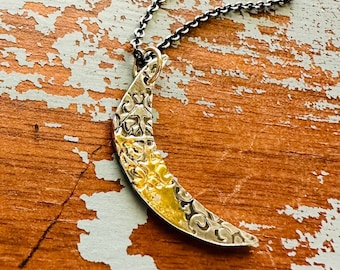 May include: A silver-toned crescent moon pendant necklace with a textured surface and a gold-colored interior. The necklace has a dark chain and rests on a distressed, painted wooden surface.