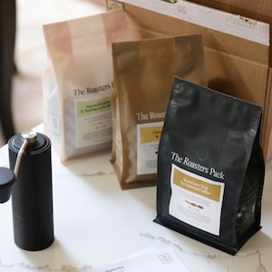 Pu&ograve; includere: Primo piano di un macinacaff&egrave; manuale nero e tre sacchetti di chicchi di caff&egrave;. I sacchetti sono etichettati "The Roasters Pack" e presentano il testo "West Coast Trail by Fernwood Coffee Company".