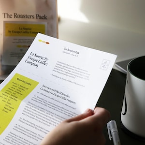 Pu&ograve; includere: Un foglio di carta bianco con il testo "La Source by Escape Coffee Company" e "The Roasters Pack" stampato su di esso. La carta &egrave; piegata a met&agrave; e ha una sezione gialla con il testo "About this coffee" stampato su di essa.