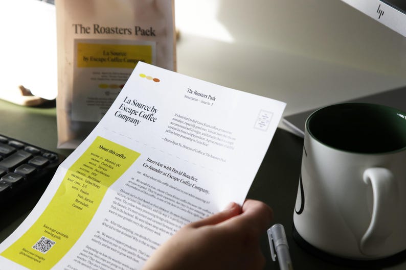 Pu&ograve; includere: Un foglio di carta bianco con il testo "La Source by Escape Coffee Company" e "The Roasters Pack" stampato su di esso. La carta &egrave; piegata a met&agrave; e ha una sezione gialla con il testo "About this coffee" stampato su di essa.