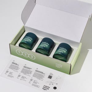 Puede incluir: Un set de degustación de matcha en una caja de cartón abierta que contiene tres latas cilíndricas de color verde oscuro etiquetadas como "matcha tasting flight". Una tarjeta informativa con descripciones numeradas, un mapa de Japón y un código QR.
