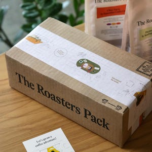 Pu&ograve; includere: Una scatola di cartone marrone con la scritta "The Roasters Pack" stampata sul lato. La scatola &egrave; appoggiata su una superficie di legno. Davanti alla scatola c'&egrave; un piccolo biglietto con la scritta "Let's go on a coffee adventure!" stampata su di esso.