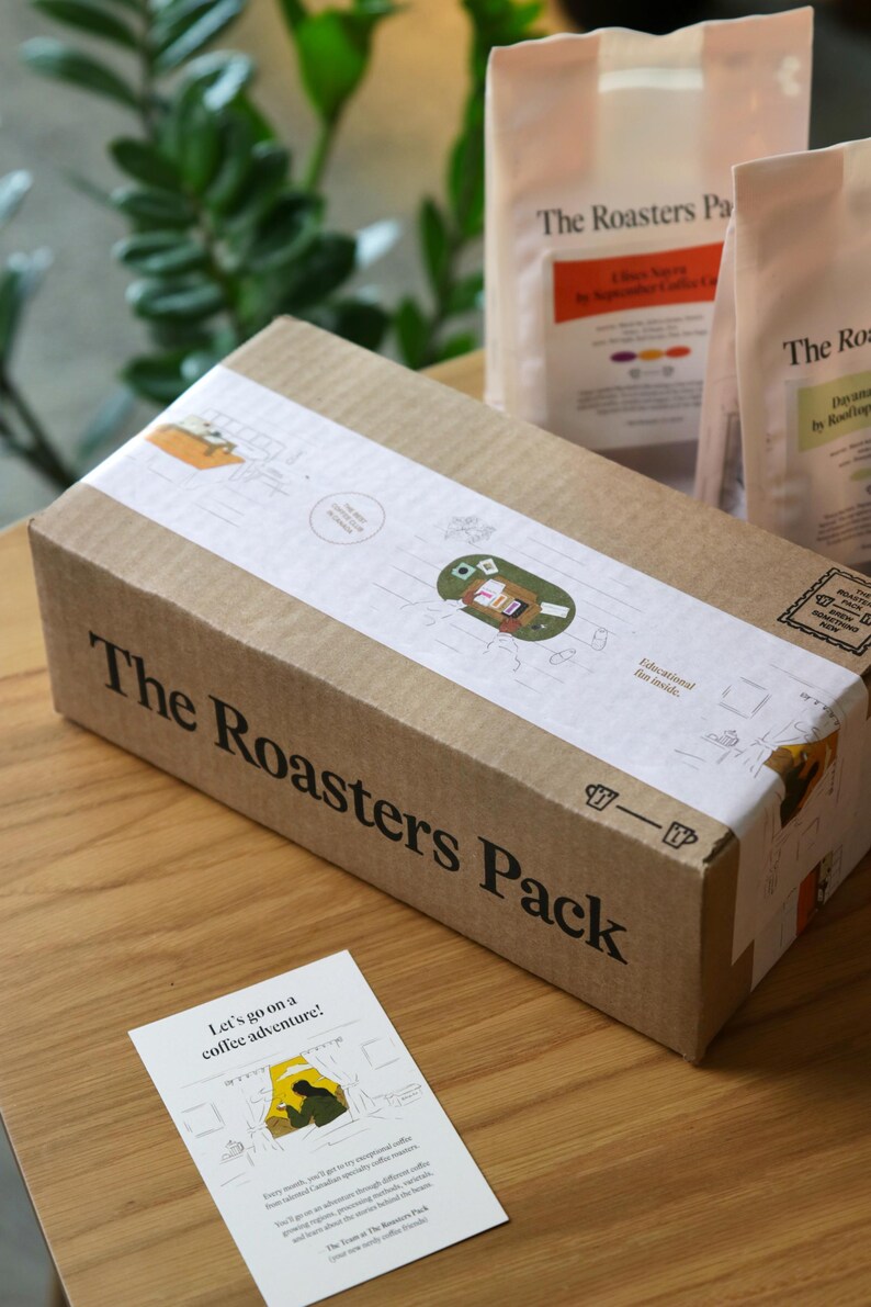 Pu&ograve; includere: Una scatola di cartone marrone con la scritta "The Roasters Pack" stampata sul lato. La scatola &egrave; appoggiata su una superficie di legno. Davanti alla scatola c'&egrave; un piccolo biglietto con la scritta "Let's go on a coffee adventure!" stampata su di esso.