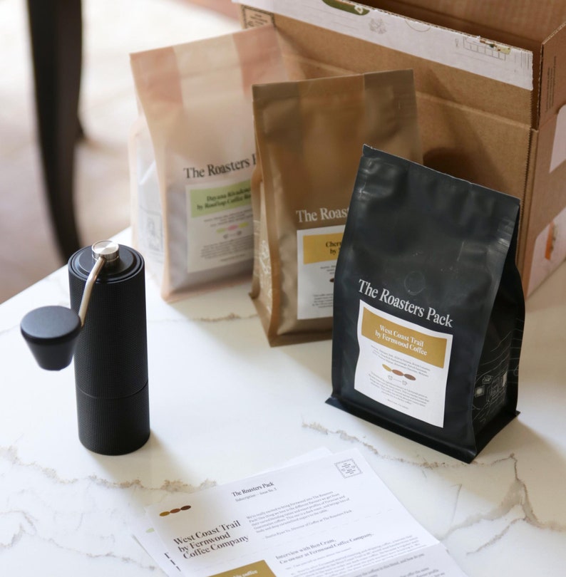 Pu&ograve; includere: Primo piano di un macinacaff&egrave; manuale nero e tre sacchetti di chicchi di caff&egrave;. I sacchetti sono etichettati "The Roasters Pack" e presentano il testo "West Coast Trail by Fernwood Coffee Company".