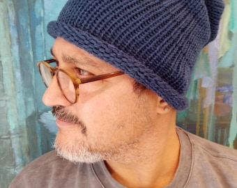 May include: A navy blue knit beanie hat with a folded brim. The hat is made of a textured, ribbed knit fabric. The person in the image is wearing glasses and a gray sweatshirt. The background is a colorful abstract painting.