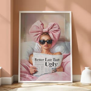 Op de afbeelding: Een ingelijste print met een vrouw met een zonnebril en een grote roze strik op haar hoofd, die een krant vasthoudt met de kop "Better Late Than Ugly". De afbeelding is geplaatst tegen een roze en witte beddengoed.