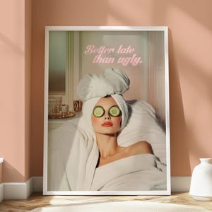 Op de afbeelding: Een ingelijste print met een vrouw in een spa-omgeving, gekleed in een witte badjas en handdoek, met komkommerschijfjes over haar ogen. De roze tekst luidt: "Better late than ugly."