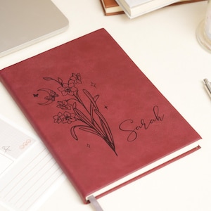 May include: A burgundy journal with a floral design and the name "Sarah" in script. The cover features a black line drawing of flowers, a crescent moon, and stars. A white pen and a small notepad are also visible.