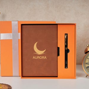 May include: An orange gift box with a brown notebook and black pen set. The notebook has the word "AURORA" and a crescent moon logo. A white ribbon wraps the box. A gold alarm clock and books are in the background.