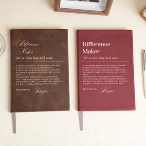 Puede incluir: Dos diarios de cuero, uno marrón y otro burdeos, cada uno con el texto "Difference Maker" y una definición. También se ven un bolígrafo, un tarro de cristales rosas y una foto enmarcada.