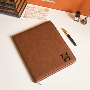 May include: A brown leather portfolio with a zipper closure. The portfolio has the name "Hamza" and the letter "H" printed in black. A black and gold pen rests on the surface next to the portfolio. Stationery.